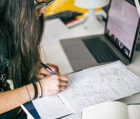 Student writing notes beside open laptop at desk